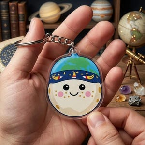 May include: A clear acrylic keychain featuring a cartoon planet design. The planet has a blue and green earth-like top, a blue headband with stars, and a smiling face with freckles. The keychain is held by a person.