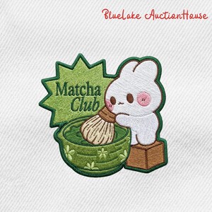 May include: Embroidered patch featuring a white bunny with pink cheeks, whisking matcha in a green bowl. The patch includes the text "Matcha Club" in green, set against a starburst design. The patch is on a white background.