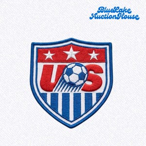 May include: A shield-shaped embroidered patch featuring the letters US in red with a soccer ball replacing the letter S. The design includes three white stars on a red background above the letters and blue and white vertical stripes below.