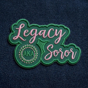 May include: Green and pink embroidered patch with the words "Legacy Soror" in cursive script. The patch includes the letters "AKA" within a circular design. The patch is on a dark blue denim background.