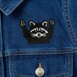 May include: A close-up of a dark blue denim jacket with a black and white raccoon patch. The patch shows a raccoon with its arms up in a peace sign. The jacket has a button closure and visible stitching.