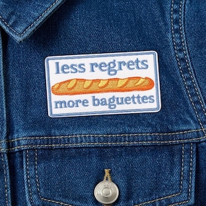 May include: A rectangular patch with the text "less regrets more baguettes" and an embroidered baguette. The patch is white with a blue border and is affixed to a blue denim jacket.