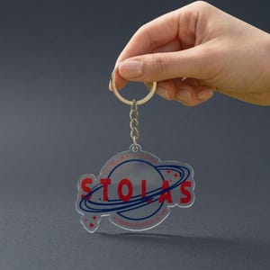 May include: Clear acrylic keychain with a silver keyring and chain. The keychain features the word "STOLAS" in red, with a blue planet and star design. Text around the planet reads "TO CALCUM AC. ASTRONOMICAL DATA COLLECTION."