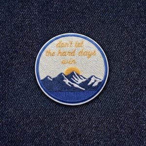 May include: Embroidered patch with a mountain landscape design. The patch features a blue and white mountain range, a yellow sun, and the text "don't let the hard days win" in gold lettering, all set against a cream background with a blue border.