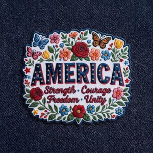 May include: Embroidered patch with the word "AMERICA" in red and blue, surrounded by colorful flowers, butterflies, and the words "Strength, Courage, Freedom, Unity". The patch has a white border and is set against a dark blue background.