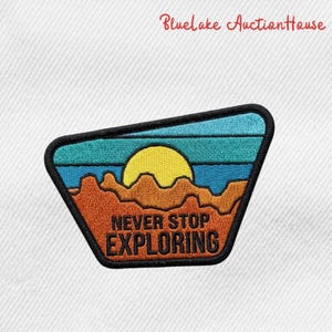 May include: Embroidered patch with a landscape design. The patch features a yellow sun, blue and teal sky, and orange mountains. The text "NEVER STOP EXPLORING" is in orange. The patch has a black border.