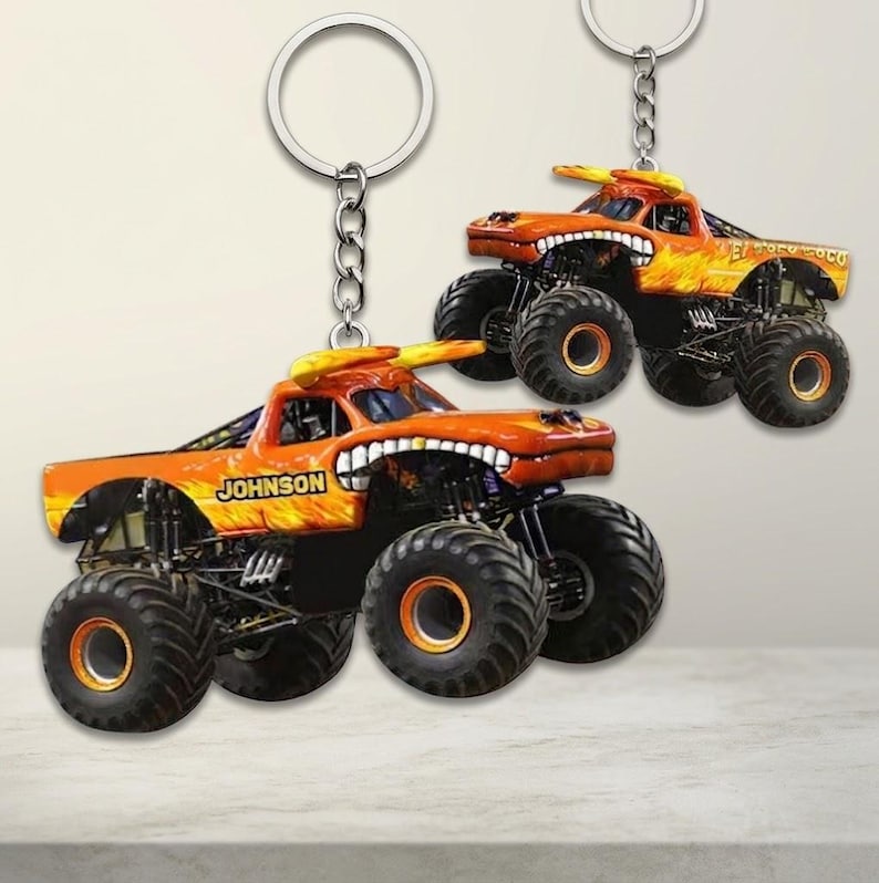 K&ouml;nnte beinhalten: Zwei orangefarbene Monster-Truck-Schl&uuml;sselanh&auml;nger mit schwarzen Reifen und einem Flammendesign. Ein Schl&uuml;sselanh&auml;nger hat den Namen "JOHNSON" auf der Seite. Der andere Schl&uuml;sselanh&auml;nger hat den Text "EL TORO LOCO". Beide haben eine silberne Kette und einen Ring.