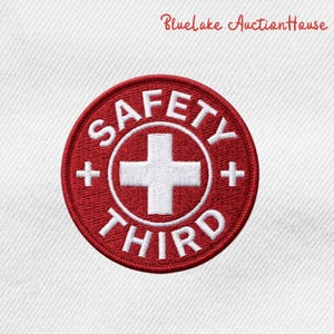 May include: Embroidered patch with a red circular design. The patch features the words "SAFETY THIRD" in white, surrounding a white cross symbol. Small white plus signs are on either side of the cross. The text "Bluelake AuctionHouse" is in the upper right corner.