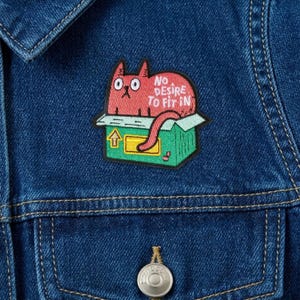 May include: A red cat patch with the text "No Desire To Fit In" is sewn onto a denim jacket. The cat is inside a green box with a yellow arrow. The patch is a fun, quirky accessory.