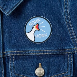 May include: A round patch featuring a white goose with a red beak and the word "HONK" on a light blue background. The patch is sewn onto a blue denim jacket.