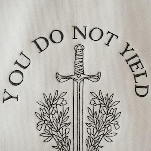 May include: White fabric with the embroidered words "YOU DO NOT YIELD" in a semi-circle above a sword and floral design. The sword is centered, with a detailed hilt and blade. Two symmetrical floral branches flank the sword.