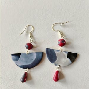 Peut inclure: Paire de boucles d'oreilles artisanales. Chaque boucle présente un motif en demi-cercle gris, noir et blanc, avec une perle rouge et grise et une perle rouge en forme de goutte. Les boucles sont suspendues à des crochets argentés.
