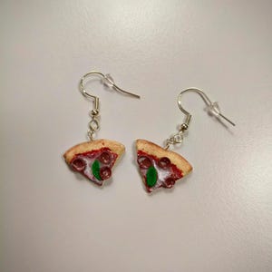 Peut inclure: Une paire de boucles d'oreilles fantaisie avec des pendentifs en forme de part de pizza. Chaque part a une croûte, de la sauce rouge, du pepperoni et une feuille de basilic verte. Les boucles d'oreilles sont suspendues à des crochets d'oreille argentés.