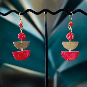 Peut inclure: Une paire de boucles d'oreilles pendantes au design géométrique. Chaque boucle d'oreille comprend un crochet doré, un petit cercle rouge, un cercle rouge plus grand avec des paillettes dorées, un demi-cercle doré et un demi-cercle rouge avec des paillettes dorées. Les boucles d'oreilles sont exposées sur un présentoir noir.