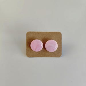 Peut inclure: Une paire de boucles d'oreilles à clous roses marbrées sur un support en carton brun. Les boucles d'oreilles rondes ont une finition brillante, avec un tourbillon de rose clair et de blanc. Un accessoire simple et élégant.