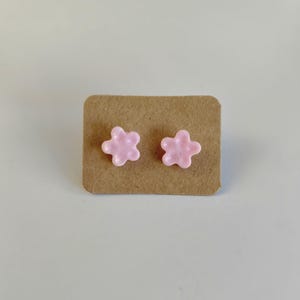 Peut inclure: Deux boucles d'oreilles clous en forme de fleur rose clair, présentées sur un support en carton brun. Les boucles d'oreilles ont une finition brillante, sur fond neutre.
