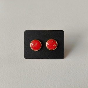 Peut inclure: Deux boucles d'oreilles rouges à clous avec des paillettes dorées et un bord doré, présentées sur un carré noir. Les boucles d'oreilles sont rondes et brillantes, sur fond neutre.