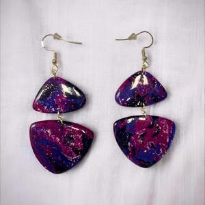 Peut inclure: Une paire de boucles d'oreilles pendantes avec un motif marbré. Chaque boucle d'oreille présente deux formes triangulaires dans les tons de violet, de bleu et d'argent. Les boucles d'oreilles ont des crochets d'oreille argentés.
