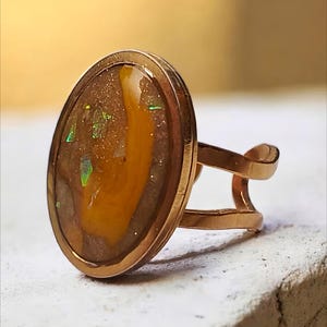 Peut inclure: Bague en or rose ornée d'une pierre ovale aux inclusions brunes, jaunes et vertes iridescentes. La bague présente un design à double bande et une finition polie, mettant en valeur la beauté naturelle de la pierre.