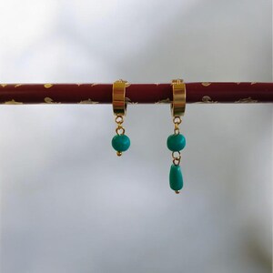 Peut inclure: Boucles d'oreilles dorées avec des perles turquoise. Une boucle a une seule perle, l'autre a deux perles rondes et une goutte. Les boucles sont suspendues sur un fond flou. Un accessoire élégant.