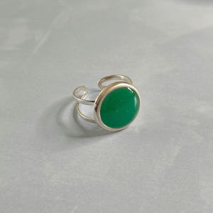 Peut inclure: Une bague réglable argentée ornée d'une pierre ronde vert émeraude. La monture de la bague présente un design à double boucle, et la pierre est sertie dans une lunette argentée polie. La bague est présentée sur une surface gris clair.