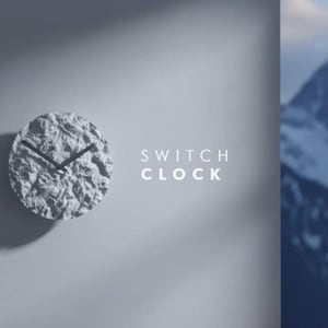 Op de afbeelding: Een ronde wandklok met een textuurrijk, wit, rotsachtig oppervlak en twee zwarte wijzers. De tekst "SWITCH CLOCK" staat in het wit rechts van de wijzerplaat gedrukt. Het ontwerp heeft een hoog contrast met diepe schaduwen.