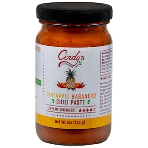 May include: A jar of Cindy's Pineapple Habanero Chili Paste. The label features a pineapple and chili peppers illustration. The jar contains a reddish-orange paste and has a black lid. Net weight is 9oz (255 g).