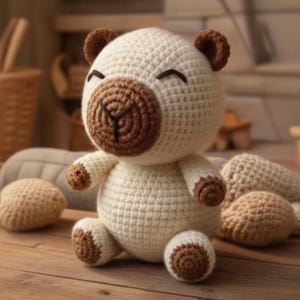 May include: A handmade crochet bear in cream and brown. The bear has a round head with small, dark brown ears and a brown snout. Its arms and legs are also detailed with brown accents. The bear is sitting on a wooden surface.