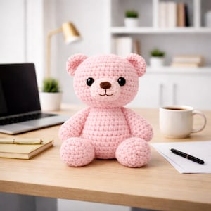 Op de afbeelding: Een handgemaakte roze gehaakte teddybeer met zwarte veiligheidsoogjes en een bruine neus. De beer zit op een houten bureau. Een laptop, koffiemok, pen en notitieblok liggen ook op het bureau.
