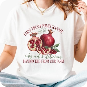 May include: White t-shirt with a pomegranate design. The graphic includes the words "FARM FRESH POMEGRANATE" and "ruby red & delicious HANDPICKED FROM OUR FARM". The shirt has a round neck and short sleeves.