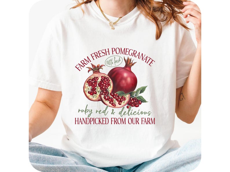 May include: White t-shirt with a pomegranate design. The graphic includes the words "FARM FRESH POMEGRANATE" and "ruby red & delicious HANDPICKED FROM OUR FARM". The shirt has a round neck and short sleeves.