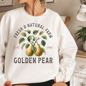 May include: A white crew neck sweatshirt with a printed design. The design features the text "Fresh & Natural Farm", a branch with two golden pears, and the words "Golden Pear". The sweatshirt also has the text "Est. 1983".