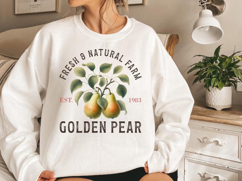 May include: A white crew neck sweatshirt with a printed design. The design features the text "Fresh & Natural Farm", a branch with two golden pears, and the words "Golden Pear". The sweatshirt also has the text "Est. 1983".