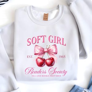 May include: White sweatshirt featuring a pink graphic with the words "SOFT GIRL" above a bow and cherries. Below the bow, it reads "Readers Society TEA AND BOOKS PROVIDED" with "1965". The sweatshirt is folded, with a tag visible.