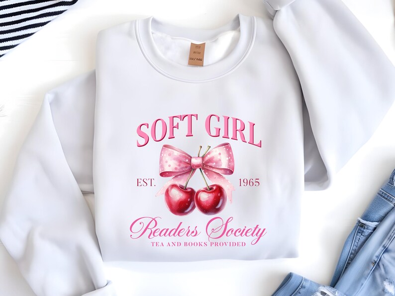 May include: White sweatshirt featuring a pink graphic with the words "SOFT GIRL" above a bow and cherries. Below the bow, it reads "Readers Society TEA AND BOOKS PROVIDED" with "1965". The sweatshirt is folded, with a tag visible.