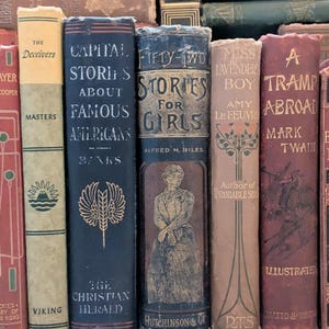 Puede incluir: Una colecci&oacute;n de libros antiguos con varios t&iacute;tulos y colores. Las lomos muestran t&iacute;tulos como "Capital Stories About Famous Americans" y "Fifty-Two Stories for Girls". Los libros est&aacute;n dispuestos verticalmente, mostrando sus cubiertas envejecidas y elementos decorativos.