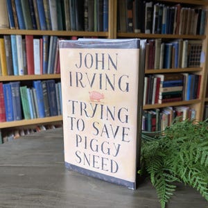 Może przedstawiać: Twarda oprawa książki "Trying to Save Piggy Sneed" autorstwa Johna Irvinga. Okładka w kolorze beżowym z czarnym tekstem i ilustracją różowej świni. Książka leży na drewnianym stole, w tle regał z książkami.