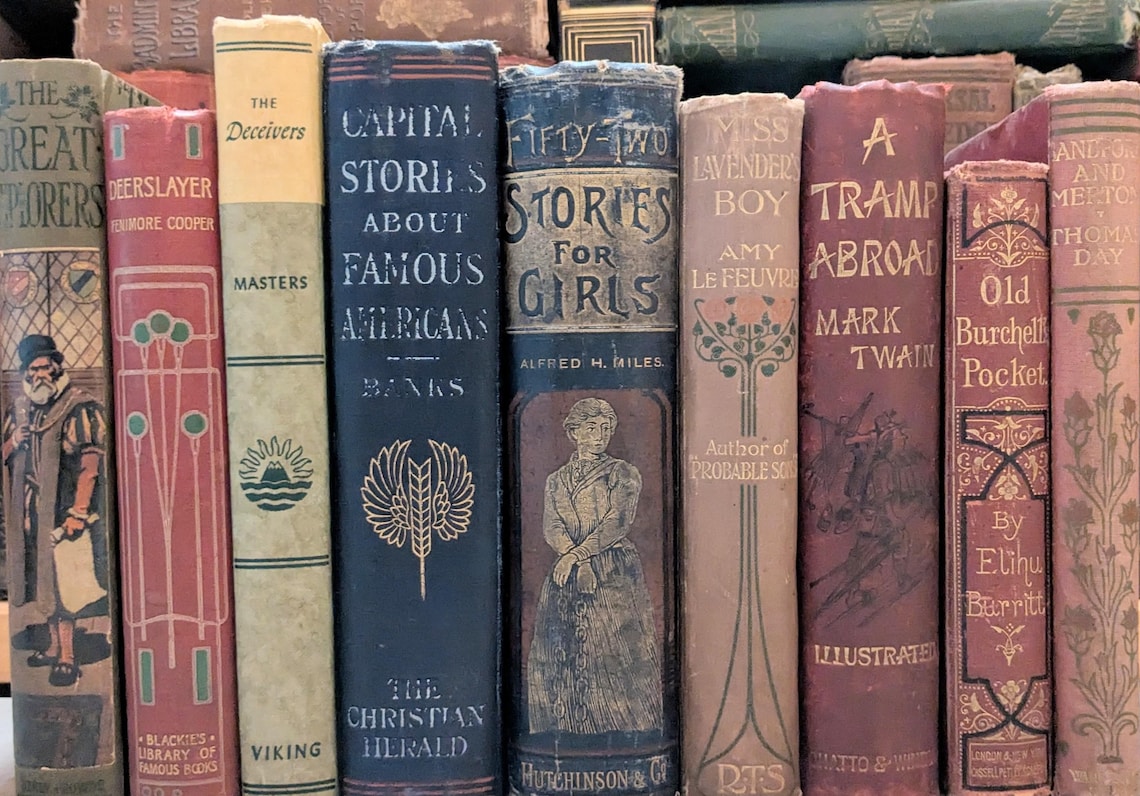 May include: A collection of antique books with various titles and colors. The spines display titles like "Capital Stories About Famous Americans" and "Fifty-Two Stories for Girls." The books are arranged vertically, showcasing their aged covers and decorative elements.