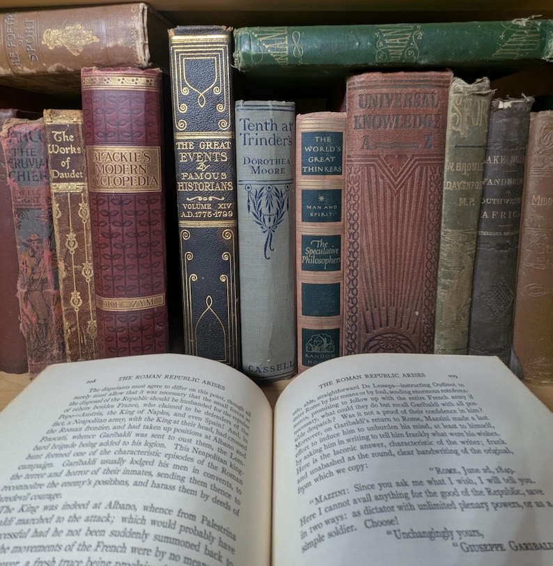 Puede incluir: Una colecci&oacute;n de libros antiguos con diversas cubiertas y dise&ntilde;os. T&iacute;tulos incluyen "The Works of Daudet" y "Blackie's Modern Encyclopedia". Un libro abierto en primer plano muestra texto.