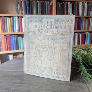 Puede incluir: Un libro antiguo titulado "The British Columbia Readers, Second Reader" con una cubierta azul claro e ilustraciones. El libro se muestra sobre una mesa de madera con un fondo de estanterías llenas de libros.