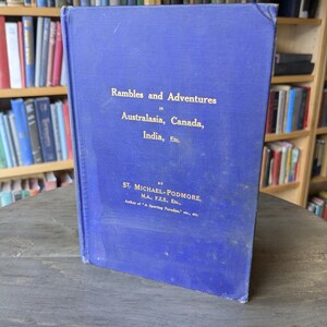 May include: A vintage blue book titled "Rambles and Adventures in Australasia, Canada, India, Etc." The cover features gold lettering and is slightly worn. The book is standing on a wooden surface, with bookshelves in the background.