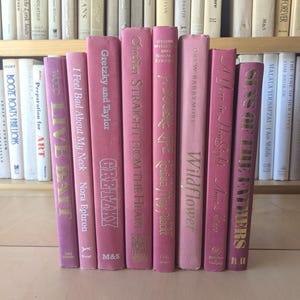 Puede incluir: Una colección de libros rosas de tapa dura con letras doradas. Los títulos incluyen "Live Bait", "I Feel Bad About My Neck", "Gretzky and Taylor" y "Sins of the Fathers". Los libros están dispuestos sobre una superficie de madera.