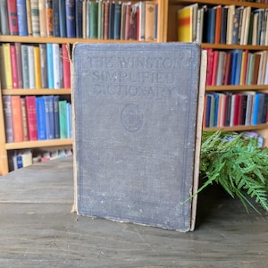 May include: An antique book titled "The Winston Simplified Dictionary" with a faded blue cover. The book is standing upright on a wooden surface, with a blurred background of bookshelves filled with books.