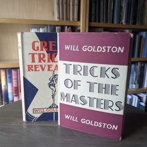 Puede incluir: Dos libros antiguos. Uno se titula "GRE TRIC REVEALED" y el otro "TRICKS OF THE MASTERS" de Will Goldston. El primer libro tiene una cubierta roja, blanca y azul con una ilustración. El segundo libro tiene una cubierta blanca y burdeos.