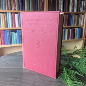 May include: A red hardcover book titled "The New Handbook of Marks and Monograms on Pottery & Porcelain" stands upright on a wooden surface. The book's cover features embossed text and a decorative emblem. A bookshelf filled with books is in the background.