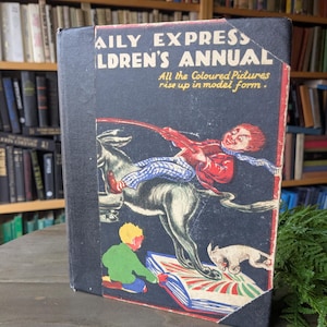May include: A vintage "Daily Express Children's Annual" book with a black cover and colorful illustrations. The cover features a boy riding a donkey, a boy reading a book, and a dog. The text reads "All the Coloured Pictures rise up in model form."