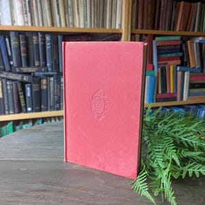 Puede incluir: Un libro de tapa dura rojo con un diseño en relieve en la portada. El libro está de pie sobre una superficie de madera, con un telón de fondo de estanterías llenas de libros. Hojas de helecho verdes están en el lado derecho del libro.