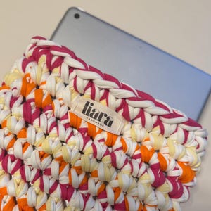 Könnte beinhalten: Eine handgefertigte Tablethülle mit einem lebendigen Häkelmuster. Die Hülle hat eine Mischung aus weißem, orangefarbenem, gelbem und magenta Stoff. Das Etikett lautet "liara handmade". Ein Tablet ist teilweise in der Hülle sichtbar.