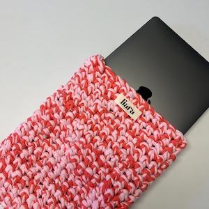 Könnte beinhalten: Eine gehäkelte Laptop-Hülle in Rosa und Rot mit einem schwarzen Laptop im Inneren. Die Hülle hat ein strukturiertes, gewebtes Aussehen und ein kleines Etikett mit der Aufschrift "liara". Die Hülle dient zum Schutz eines Laptops.