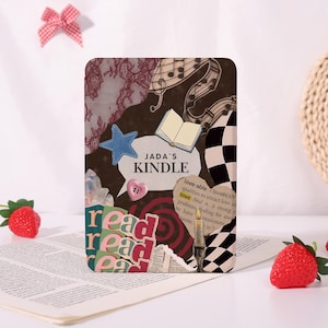 May include: A decorative Kindle cover with a collage design. The cover features a blue star, a pink heart-shaped button, and the text "JADA'S KINDLE". Other elements include musical notes, a checkered pattern, and the word "read".